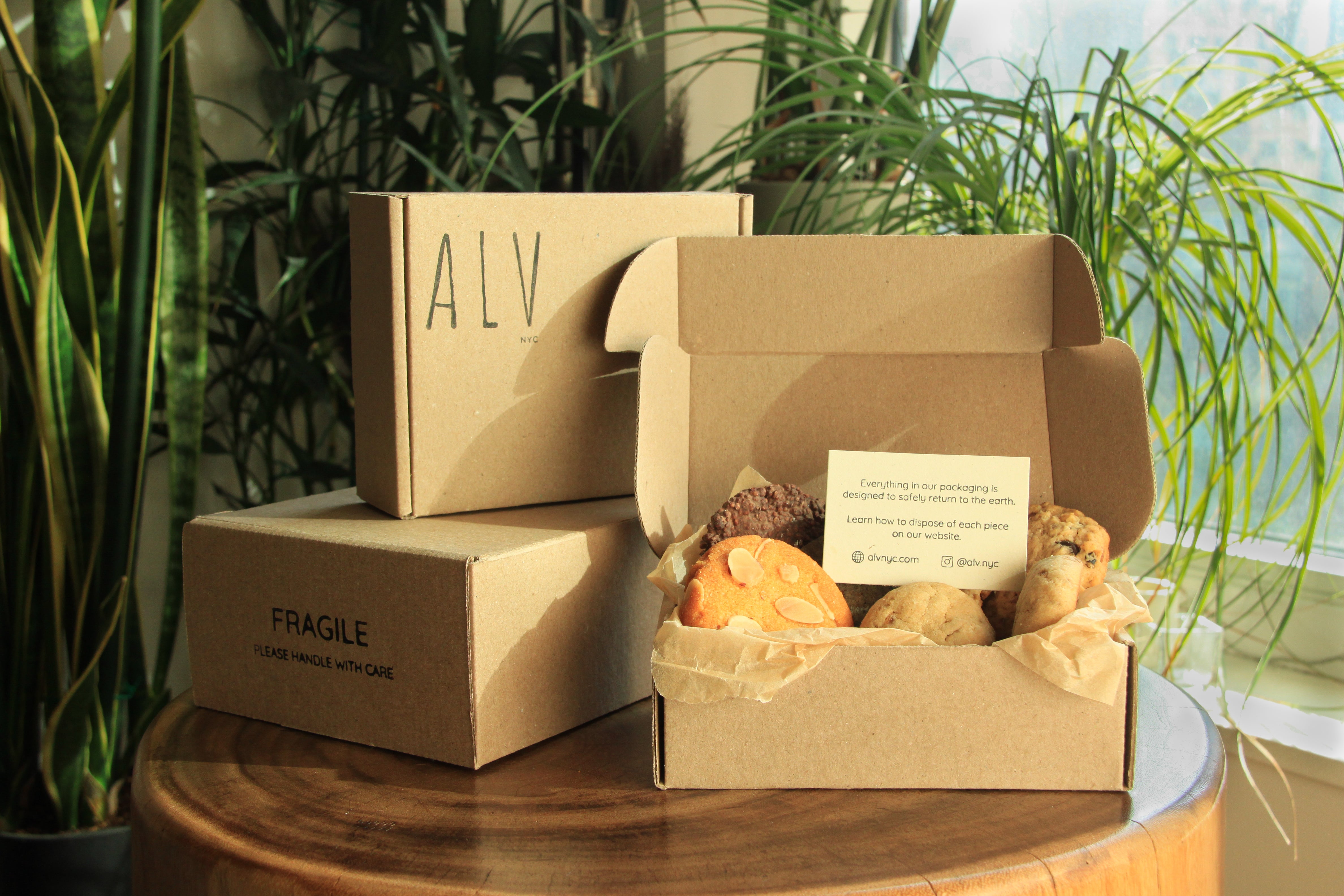 ALV NYC cookie delivery boxes open and closed, showing compostable kraft paper packaging, tissue paper lining, and insert card on a wooden table surrounded by plants